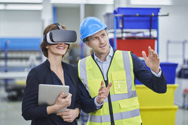 Supervisor explaining plan to female manager wearing VR glasses while holding digital tablet in factory - RORF02165