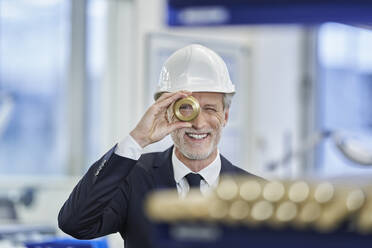 Happy male manager in white hardhat looking through brass object in factory - RORF02210