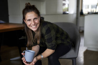 Lächelnde Frau, die eine Kaffeetasse hält und wegschaut, während sie auf einem Stuhl zu Hause sitzt - AUF00549