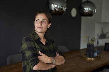 Thoughtful woman with arms crossed looking away while standing by dining table at home - AUF00552