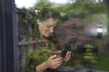 Lächelnde Frau im mittleren Erwachsenenalter, die zu Hause ein Smartphone benutzt, gesehen durch ein Fenster - AUF00553