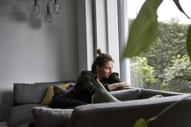 Nachdenkliche Frau schaut durch das Fenster, während sie sich zu Hause auf dem Sofa entspannt - AUF00560