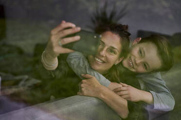 Mutter nimmt Selfie mit Sohn beim Entspannen auf dem Sofa zu Hause durch das Fenster gesehen - AUF00574