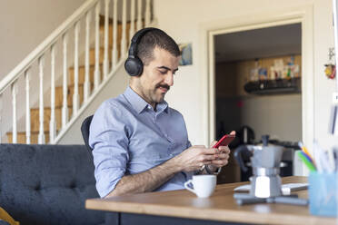 Man working from home, using smartphone with headphones during coffee break - WPEF02980