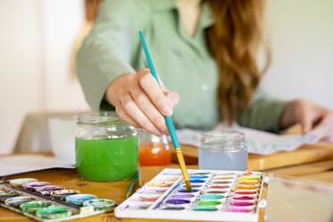 Young woman dipping paintbrush in watercolor on table at home - AFVF06495