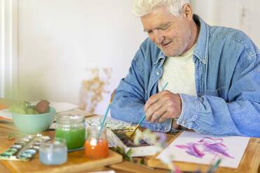 Glücklicher älterer Mann malt mit Aquarellfarben auf dem Tisch zu Hause - AFVF06497