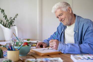 Glücklicher älterer Mann malt auf Papier am Tisch - AFVF06514