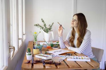 Nachdenkliche junge Frau hält Pinsel beim Malen auf dem Tisch zu Hause - AFVF06517