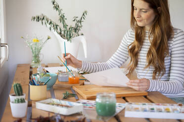 Young woman dipping paintbrush in water while painting at home - AFVF06518