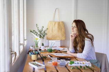 Thoughtful young woman looking away while painting on table at home - AFVF06523