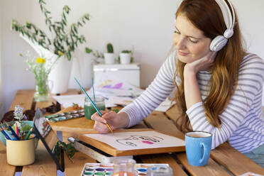 Young woman listening music though headphones while painting at home - AFVF06529