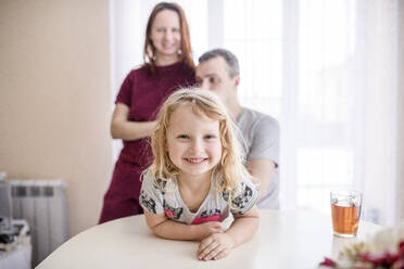 Glückliches süßes Mädchen lehnt sich an Esstisch mit Eltern stehen im Hintergrund zu Hause - EYAF01117