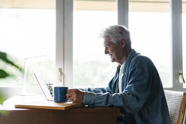 Happy senior man holding coffee mug while using laptop at home - AFVF06539