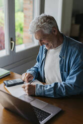 Happy senior man writing on paper while using calculator and laptop at home - AFVF06543
