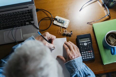 Ausgeschnittenes Bild eines älteren Mannes, der bei der Reparatur eines Laptops zu Hause ein Diagramm auf Papier erstellt, lizenzfreies Stockfoto