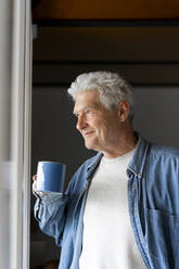 Nachdenklicher älterer Mann lächelt beim Kaffeetrinken am Fenster zu Hause - AFVF06552