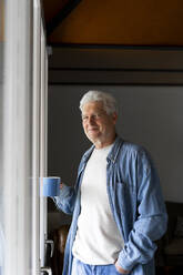 Selbstbewusster älterer Mann, der eine Kaffeetasse hält, während er zu Hause am Fenster steht - AFVF06553