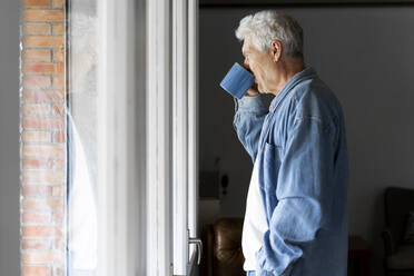 Nachdenklicher älterer Mann trinkt Kaffee, während er zu Hause am Fenster steht - AFVF06554