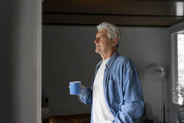 Nachdenklicher älterer Mann, der eine Kaffeetasse in der Hand hält und durch ein Fenster in seinem Haus schaut - AFVF06555