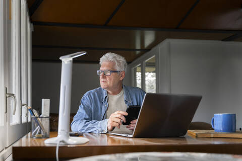 Nachdenklicher älterer Mann hält Smartphone mit Laptop auf dem Tisch zu Hause, lizenzfreies Stockfoto