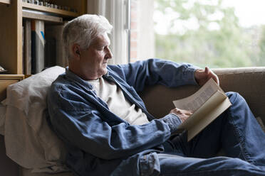 Senior man reading book while relaxing on sofa at home - AFVF06560
