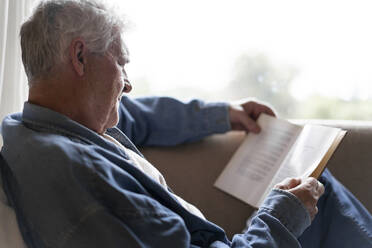 Senior man reading book while relaxing on sofa at home - AFVF06562