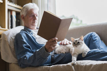 Senior man reading book while cat lying on lap at home - AFVF06566