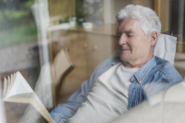 Senior man reading book while relaxing on sofa seen through glass window - AFVF06567