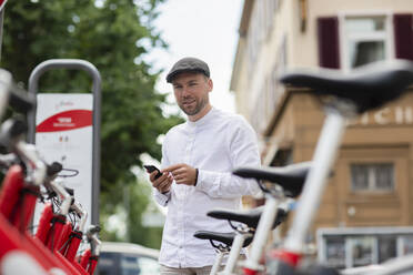 Lächelnder Mann, der über sein Smartphone an einer Parkstation in der Stadt ein Fahrrad mietet - SGF02658