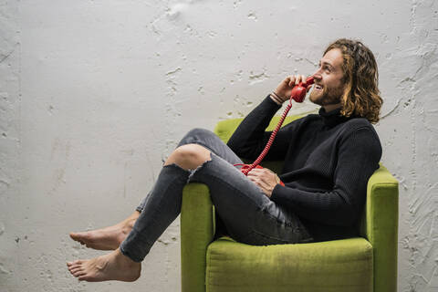 Smiling man talking over telephone while sitting on armchair against wall at home stock photo