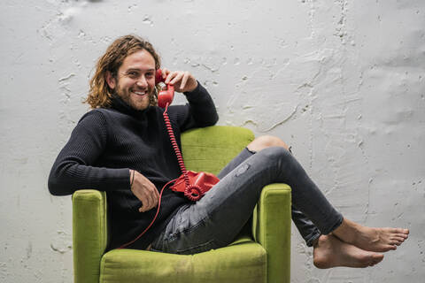 Lächelnder Mann spricht am Telefon und entspannt sich auf einem Sessel an der Wand, lizenzfreies Stockfoto