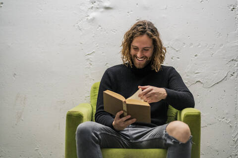 Lächelnder junger Mann liest ein Buch, während er zu Hause auf einem Sessel an der Wand sitzt, lizenzfreies Stockfoto
