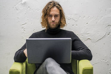 Bärtiger junger Mann, der einen Laptop benutzt, während er zu Hause auf einem Sessel an der Wand sitzt - DLTSF00725
