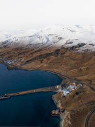 Luftaufnahme der schneebedeckten Gipfel des Berges Brekkukambur, der Bauernhäuser und Fischerboote in der Bucht und der kurvenreichen Straße um den Hvalfjordur-Fjord in Westisland. - AAEF08770