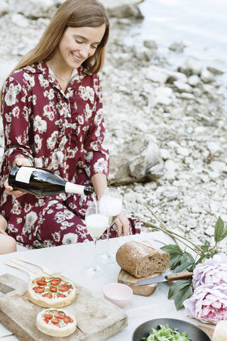 Lächelnde junge Frau gießt Wein in ein Glas, während sie am Seeufer sitzt, lizenzfreies Stockfoto