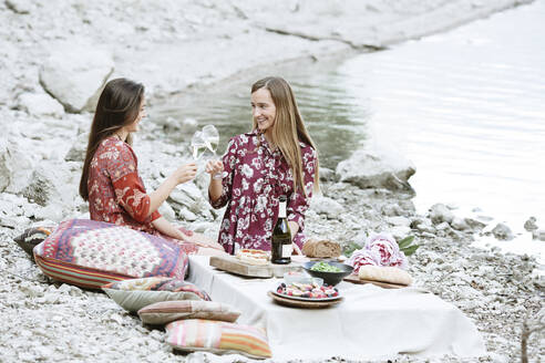 Freundinnen stoßen mit Weingläsern an, während sie am Seeufer sitzen - ALBF01248