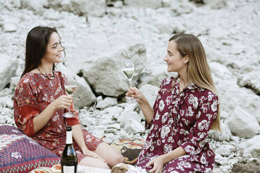 Female friends holding wineglasses talking while sitting against rocks - ALBF01249
