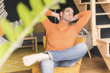 Mature man relaxing while sitting with hands behind head and eyes closed in living room at home - UUF20438