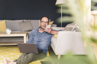 Geschäftsmann, der mit einem Mobiltelefon spricht, während er zu Hause mit einem Laptop arbeitet - UUF20447