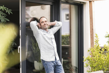 Smiling mature man leaning on window with hands behind head at back yard - UUF20449