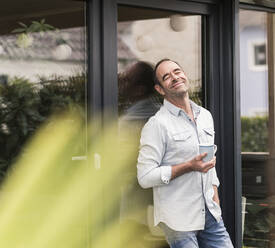 Happy mature man holding coffee cup while leaning against window at back yard - UUF20452