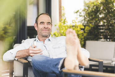 Smiling man sitting with coffee cup at back yard - UUF20454