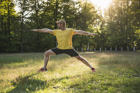 Man practicing warrior 2 position on grass against trees at park - UUF20486