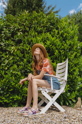 Schöne Frau, die sich auf einem Stuhl sitzend vor Pflanzen im Hinterhof entspannt - AFVF06575
