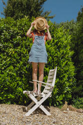 Young woman covering face with hat while standing on chair against plants at back yard - AFVF06577