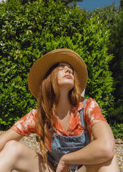 Schöne rothaarige Frau mit geschlossenen Augen sitzen gegen Pflanzen im Hinterhof auf sonnigen Tag - AFVF06578