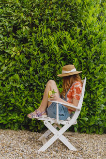 Young woman sitting with mojito cocktail on chair by green plants at back yard - AFVF06579