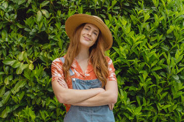 Smiling beautiful young redhead woman standing with arms crossed against green plants at back yard - AFVF06581
