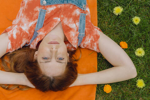 Relaxed young woman lying with hands behind head on picnic blanket by flowers at back yard stock photo