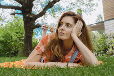 Thoughtful young redhead woman lying on picnic blanket while looking away at back yard - AFVF06587